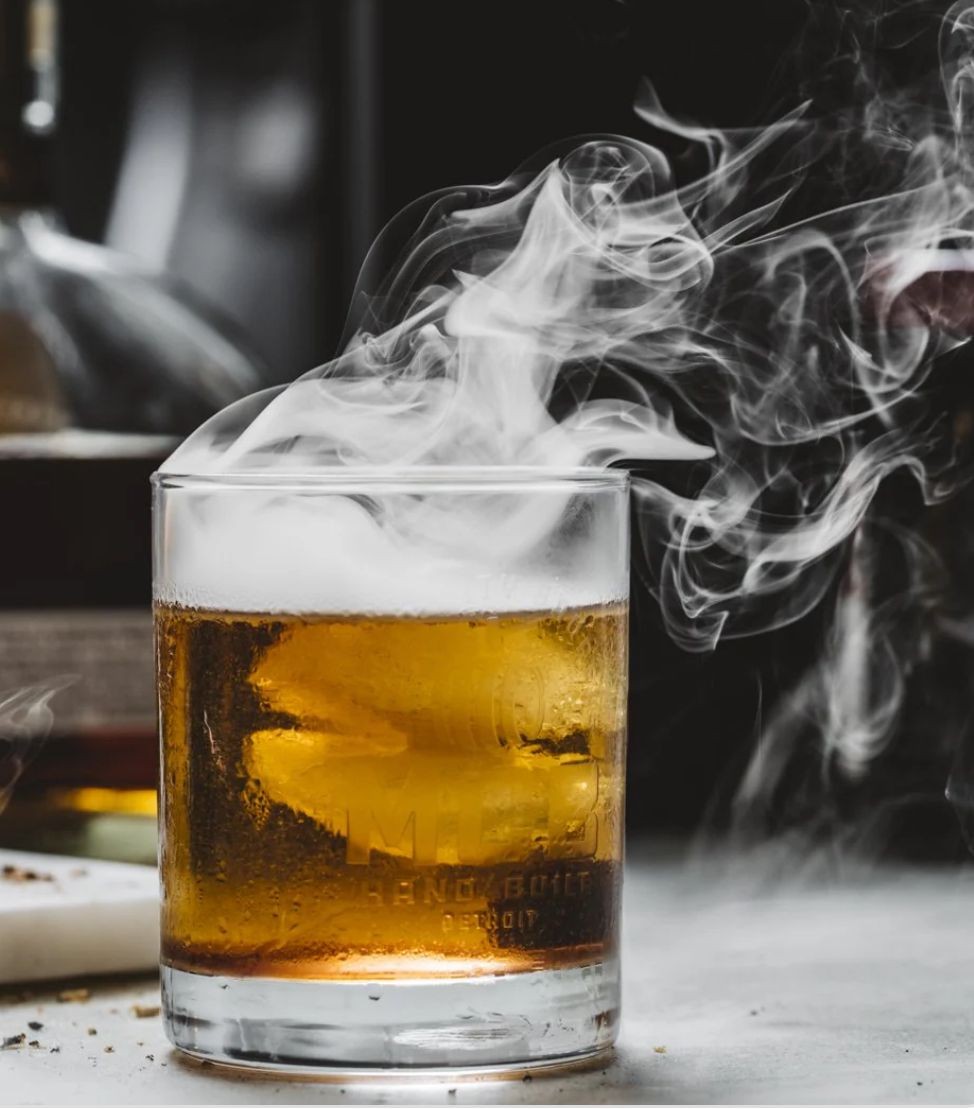 A whisky glass filled with amber liquid, emitting wisps of smoke on a dark background.