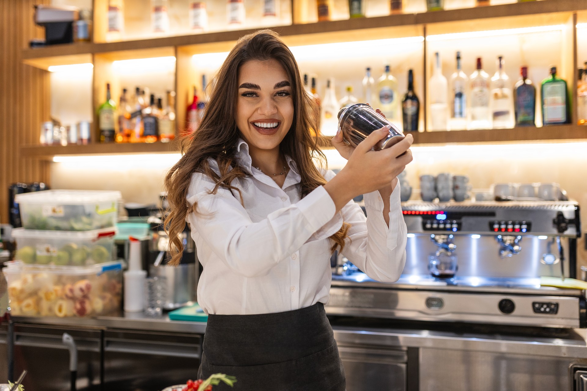 Young female worker at bartender desk in restaurant bar preparing cocktails with shaker. Beautiful young woman behind bar