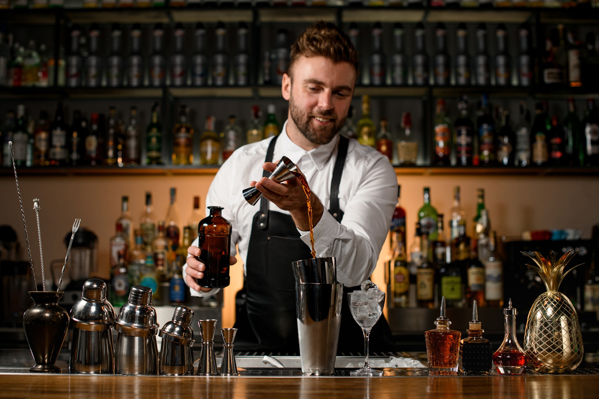 various shakers and bottles stand on the bar counter, and bartender gently pours alcoholic drink into cup