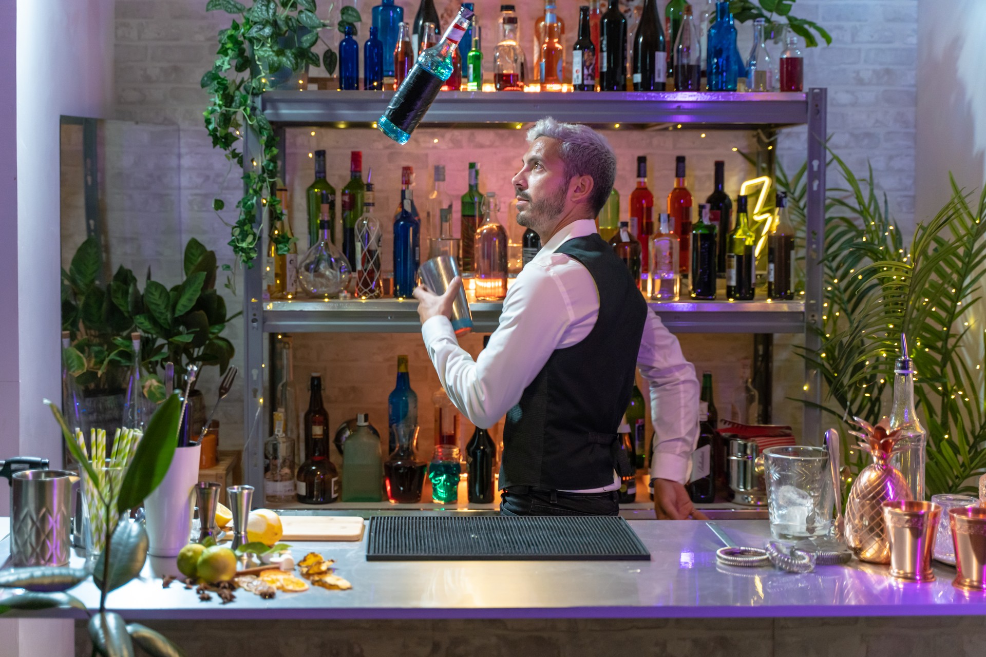 Handsome young bartender inside the bar skillfully throws a shaker with ice and a bottle into the air. Shelves with bottles on blurred background.